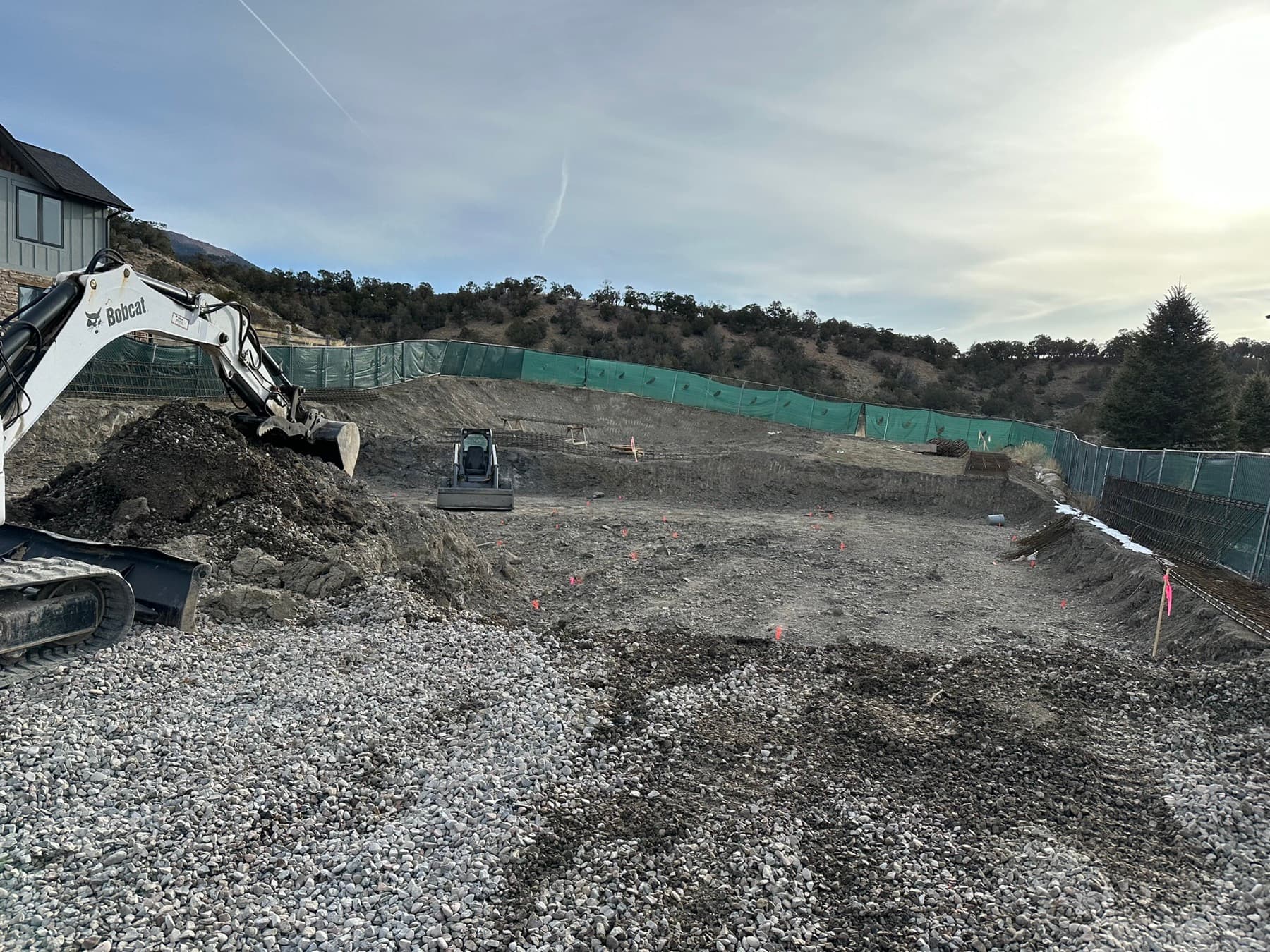 Excavator grading a home site pad with rebar footings staked out, mountain backdrop